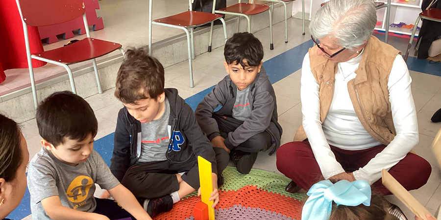 Três meninos sentados brincando com uma senhora idosa dentro da sala de aula